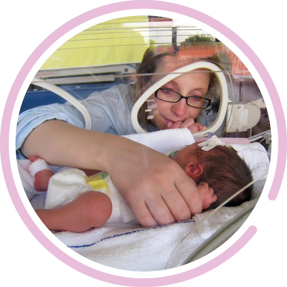 New mother touching her premature newborn's hand while they rest in a NICU incubator.